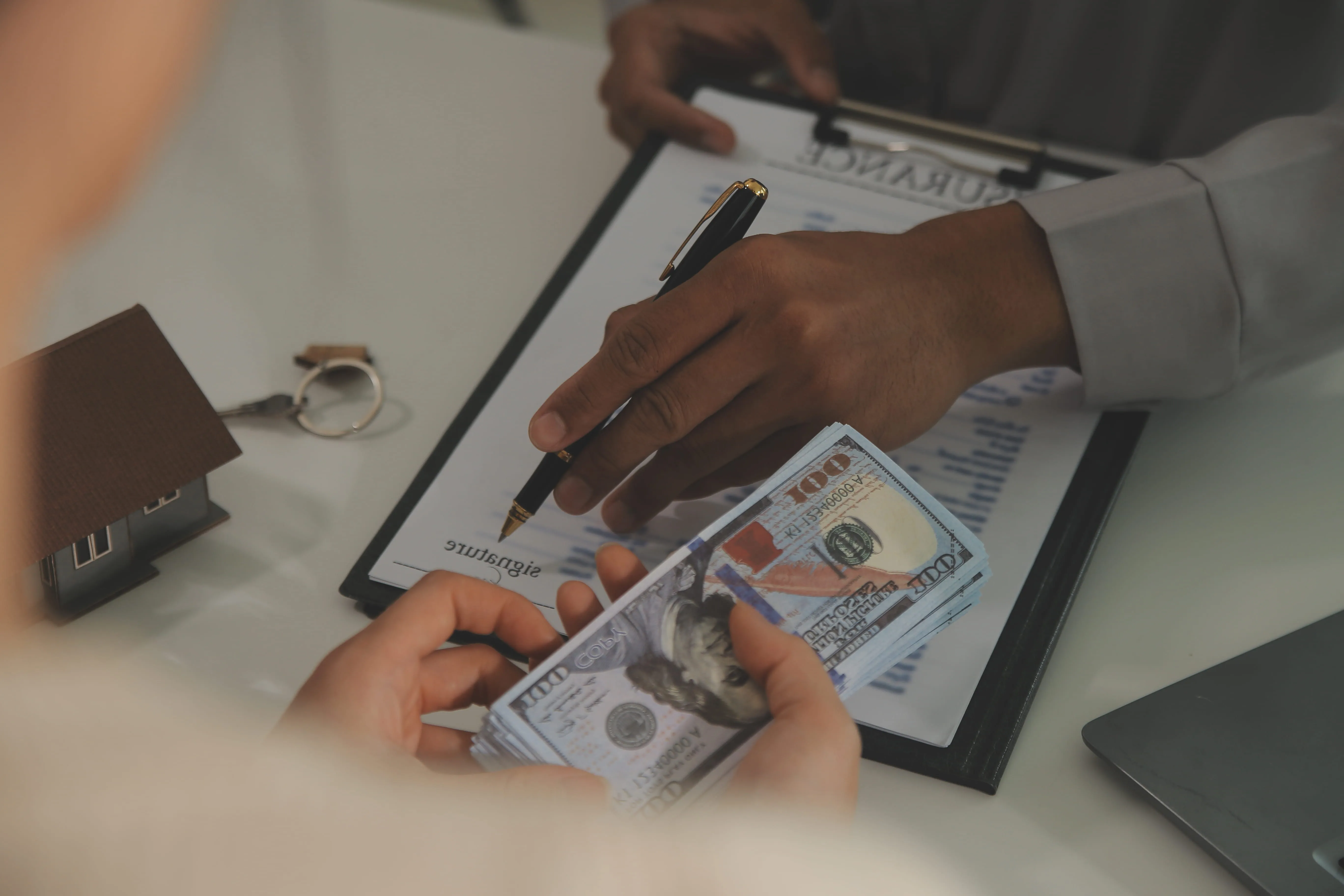 Person signing a contract while another holds a stack of $100 bills near a house key and laptop.