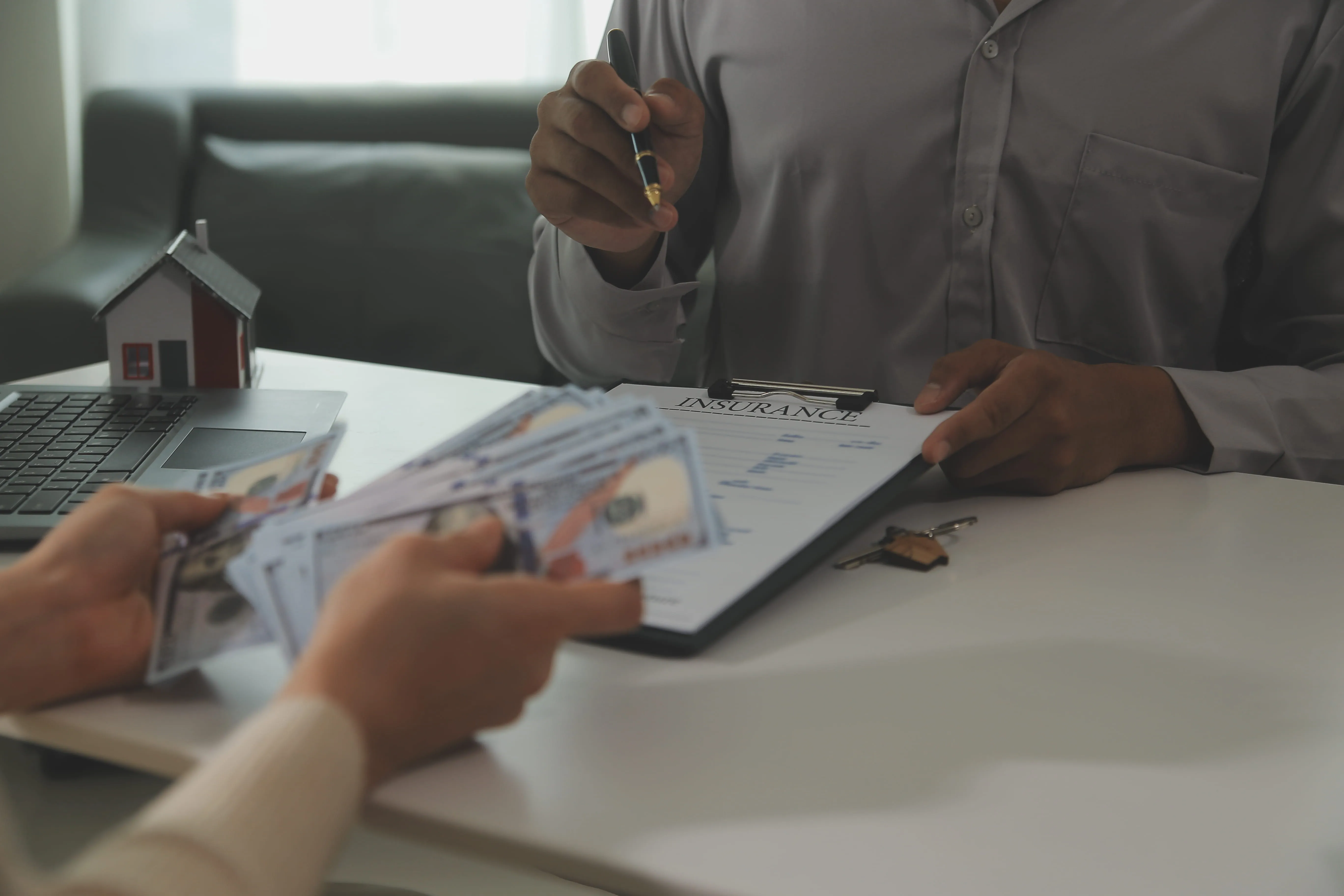 Person holding money with another showing insurance document and pen over a table with house model and keys.