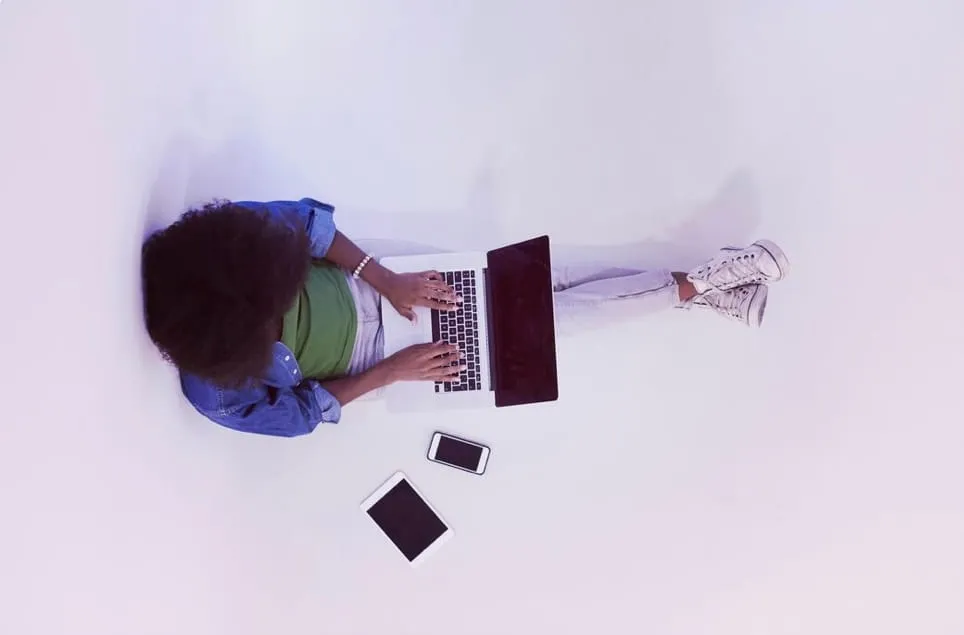 Top view of person in denim jacket typing on laptop with tablet and smartphone nearby on white floor.