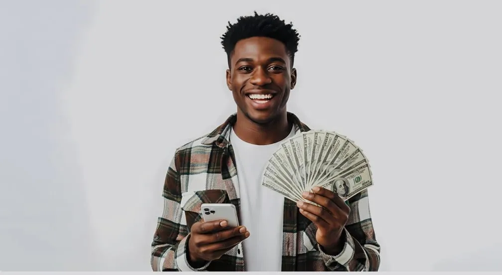 Smiling young man in plaid shirt holding a smartphone and fanned US hundred-dollar bills.