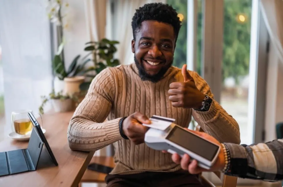 Man in a beige sweater pays by card with a terminal and gives a thumbs-up, smiling.