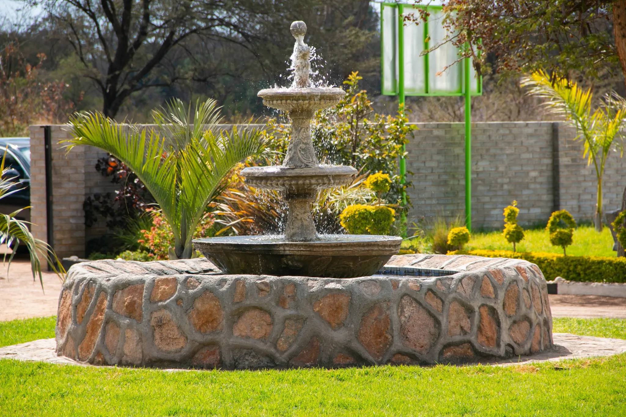 Stone three-tier garden fountain with water splashing, surrounded by greenery and plants.