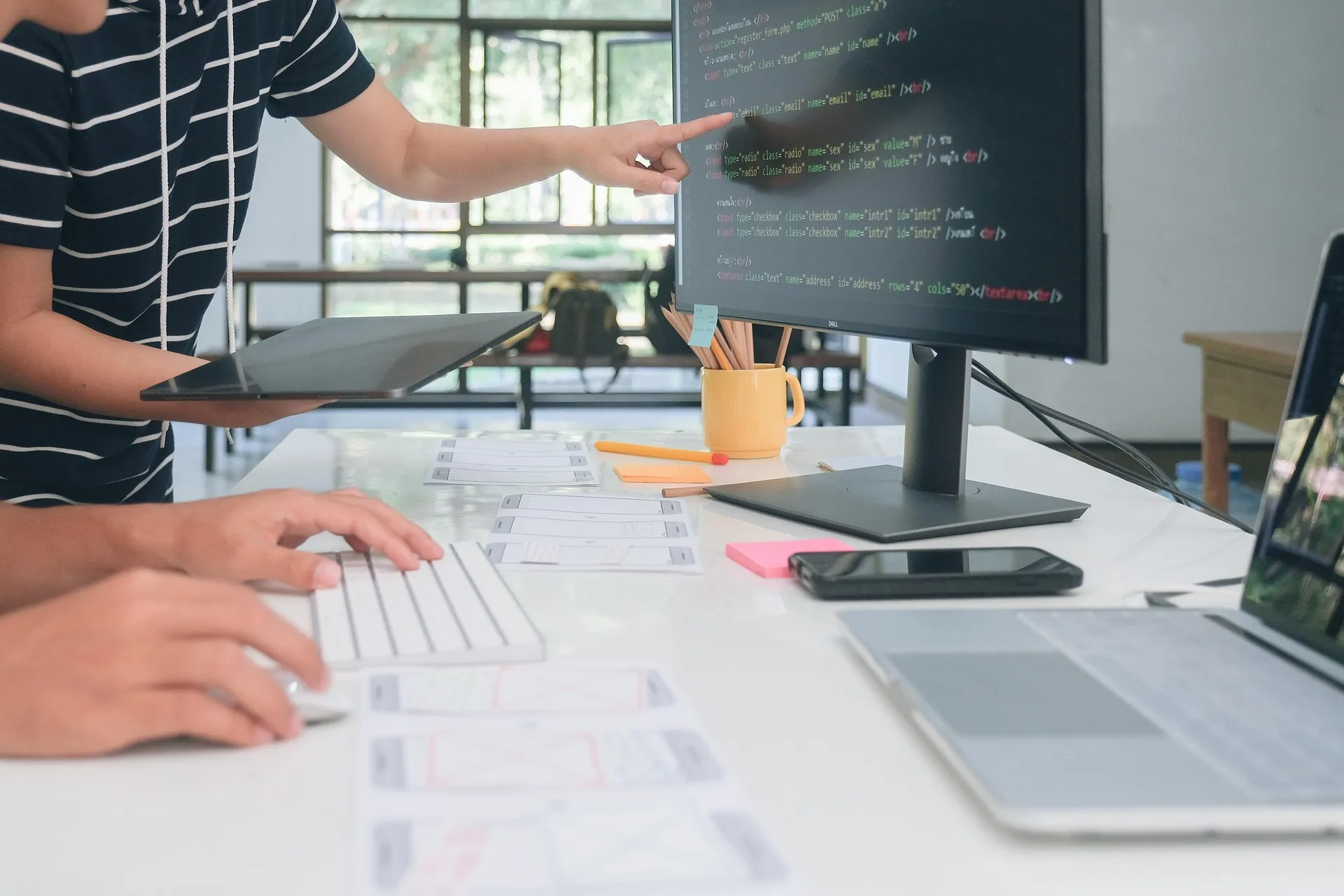 Person pointing at computer screen with code while another types on keyboard at a workspace.
