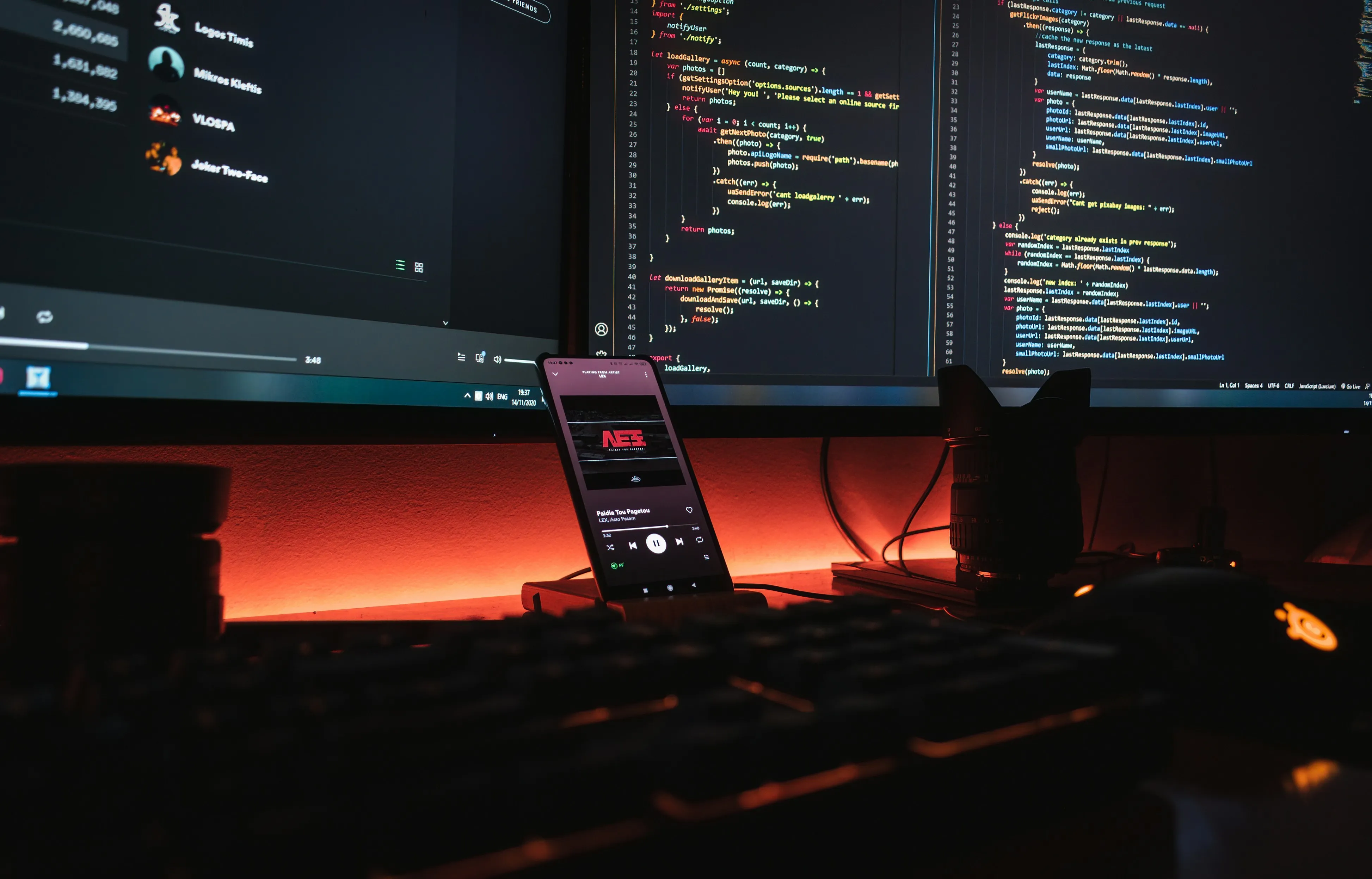 Desk setup with dual monitors showing code, smartphone playing music, keyboard, camera lens, and mouse lit orange.