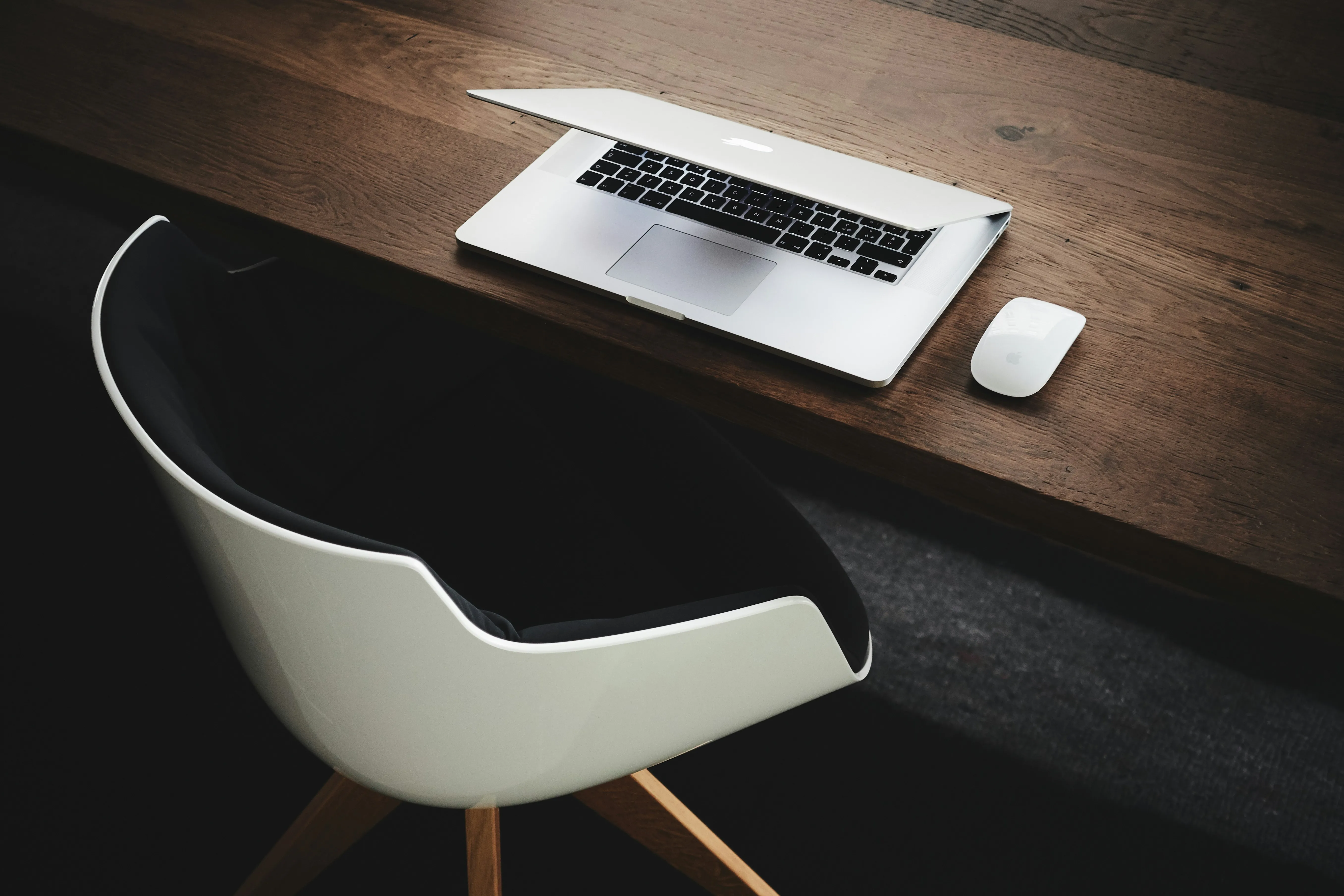 Partially open laptop and wireless mouse on a wooden desk beside a modern white chair.