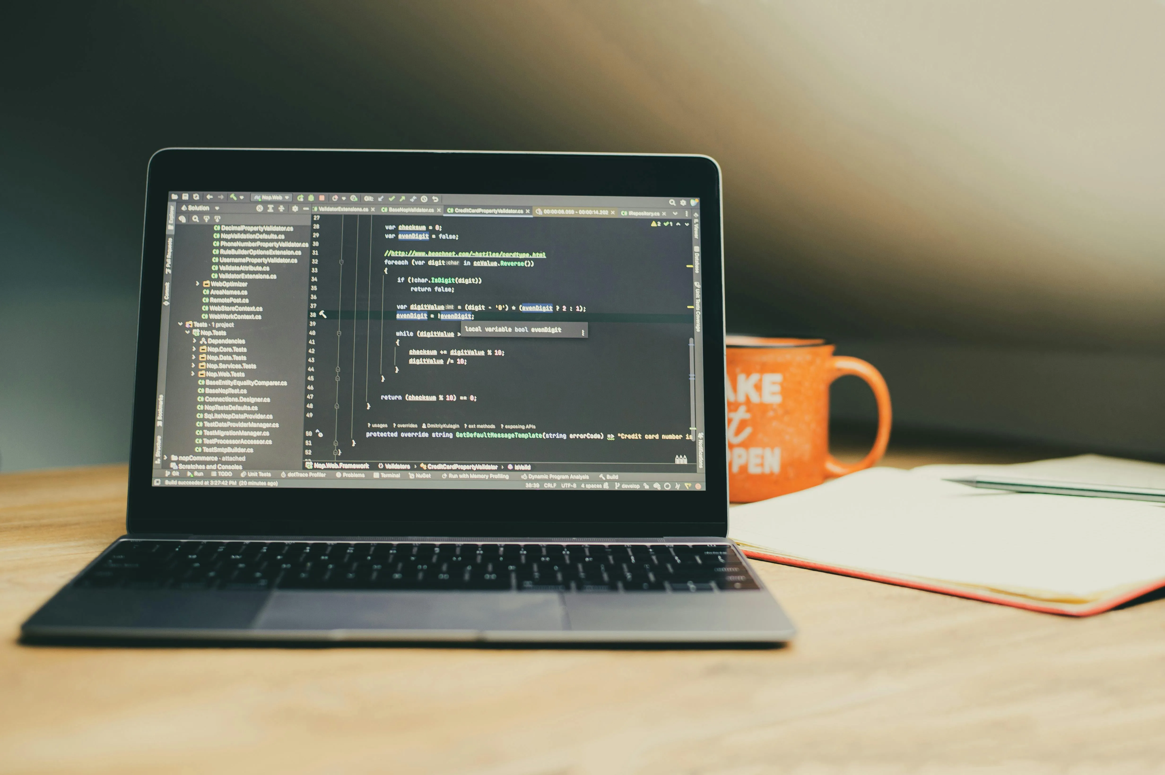 Laptop on wooden desk displaying code editor with programming code, next to orange coffee mug and open notebook.