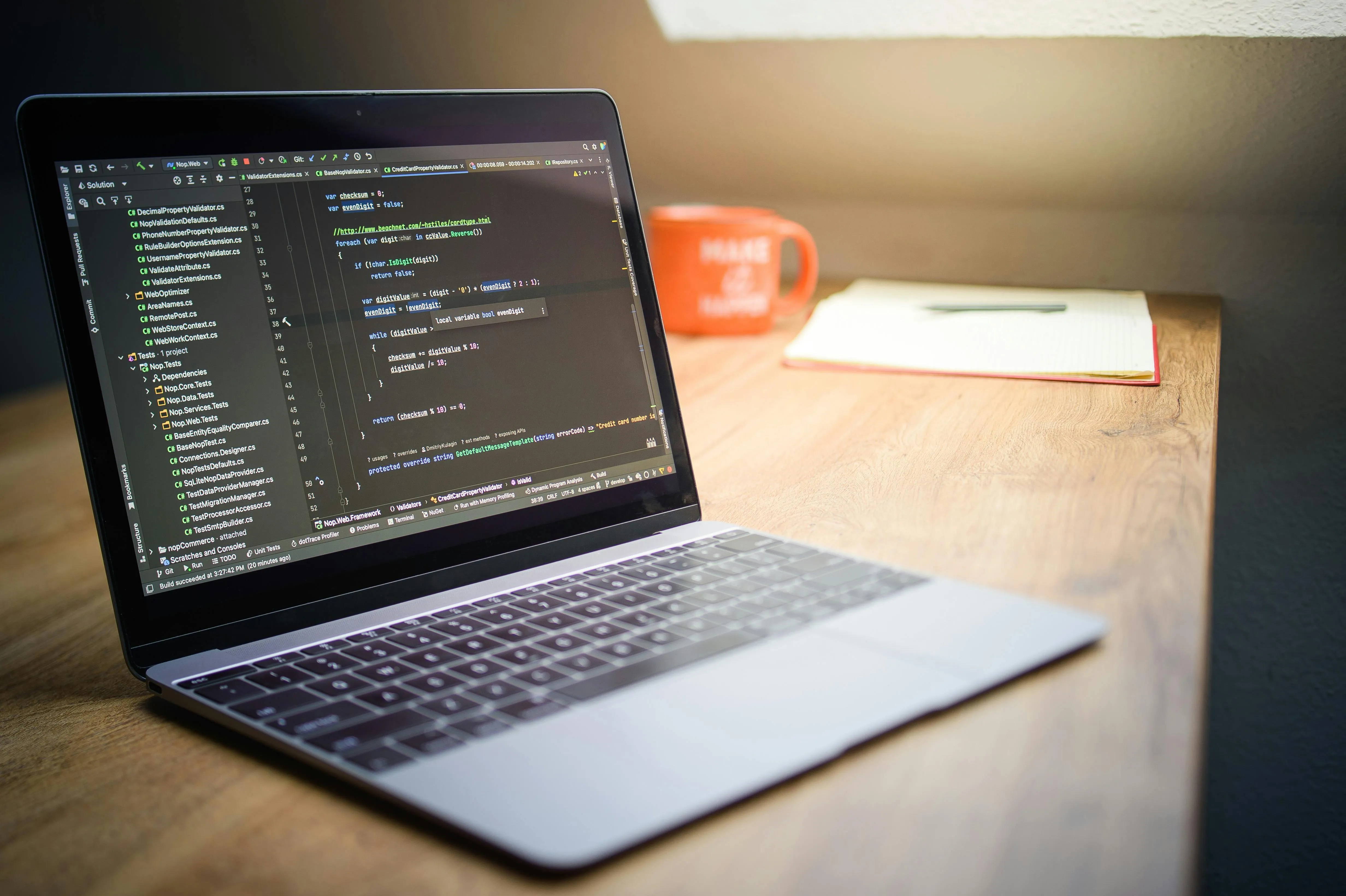 Laptop displaying source code on a wooden desk with a blurred orange mug and notebook in background.