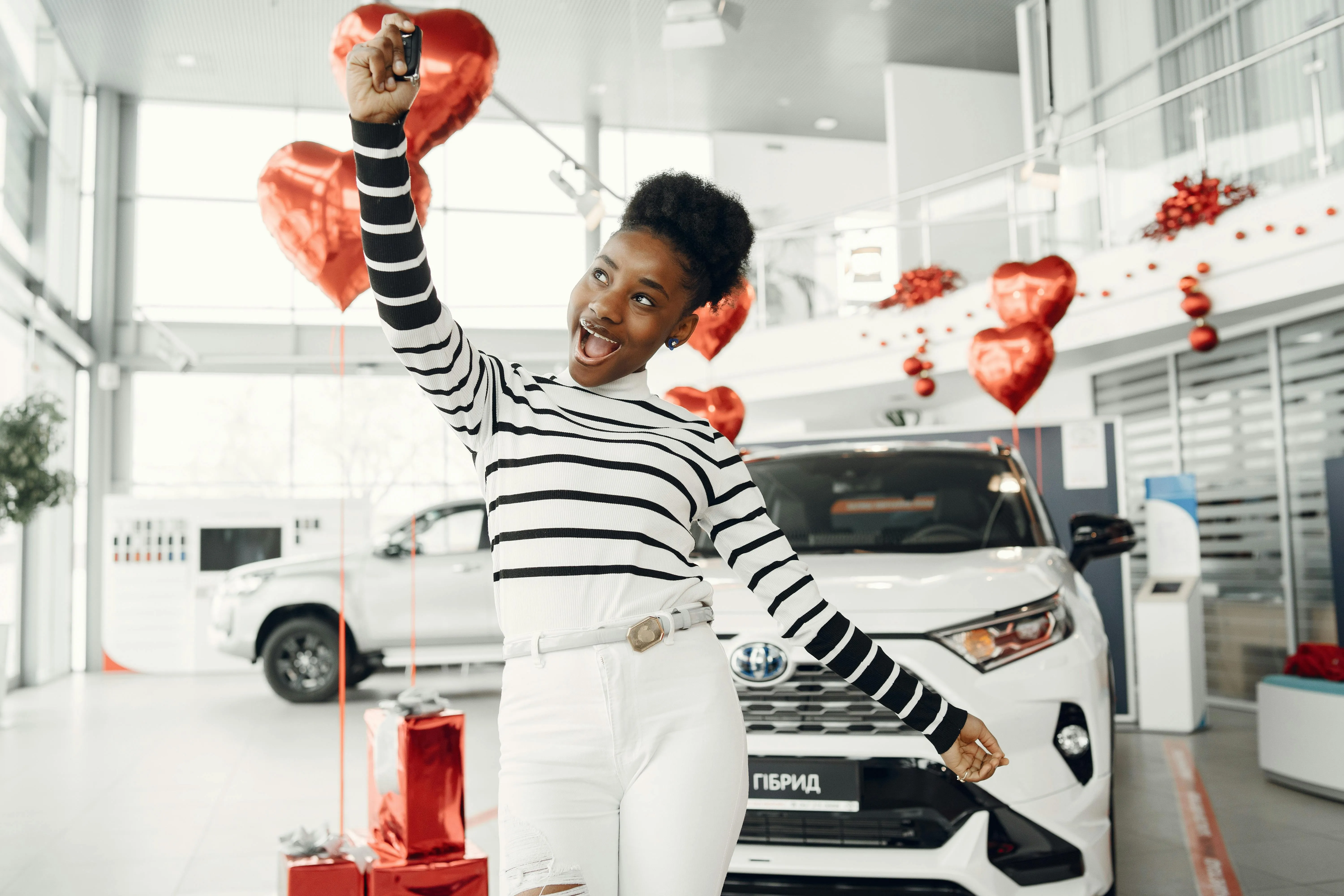 Excited woman holding up car keys in front of a white SUV inside a decorated showroom.