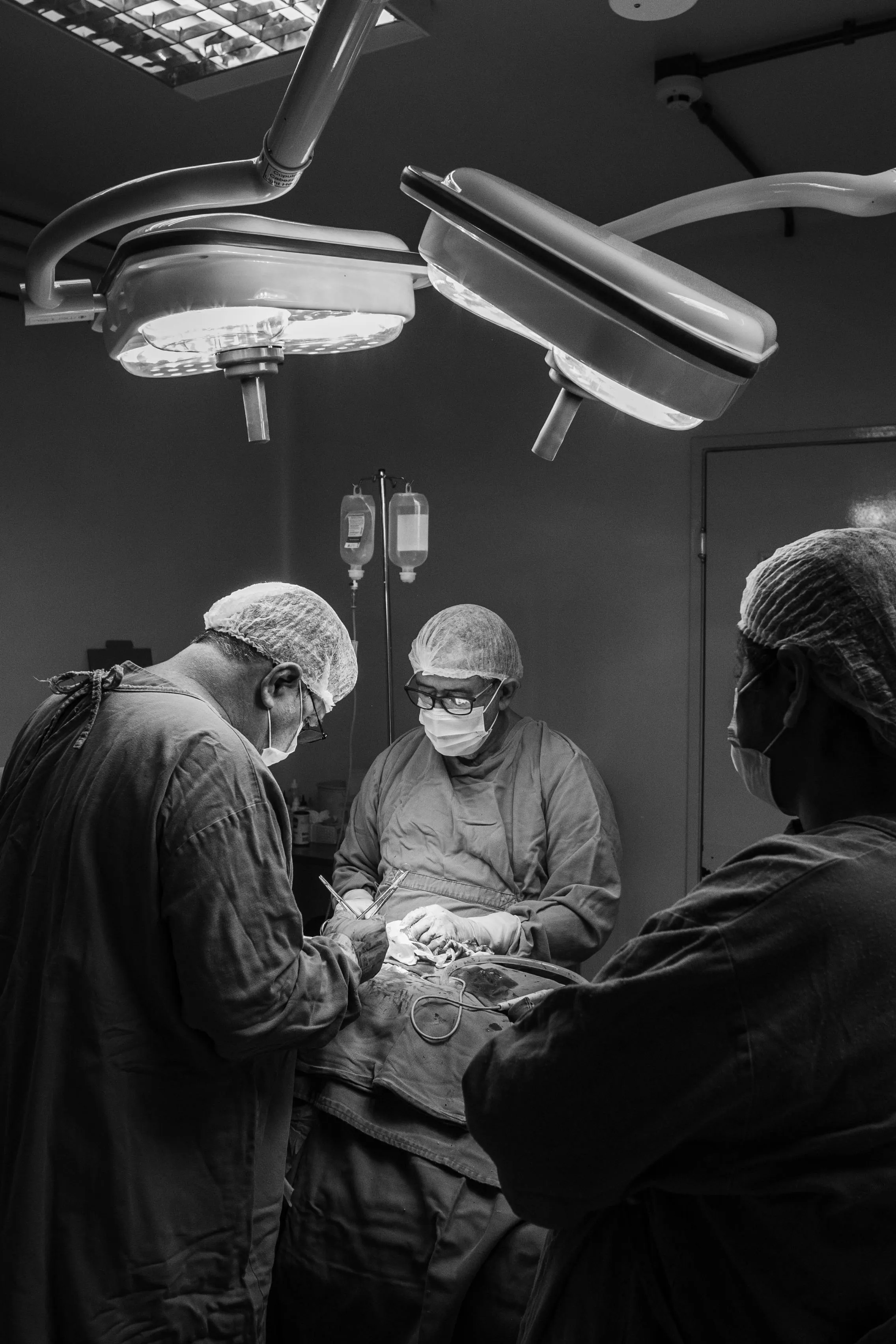 Three surgeons wearing masks and caps performing surgery under bright operating lights.