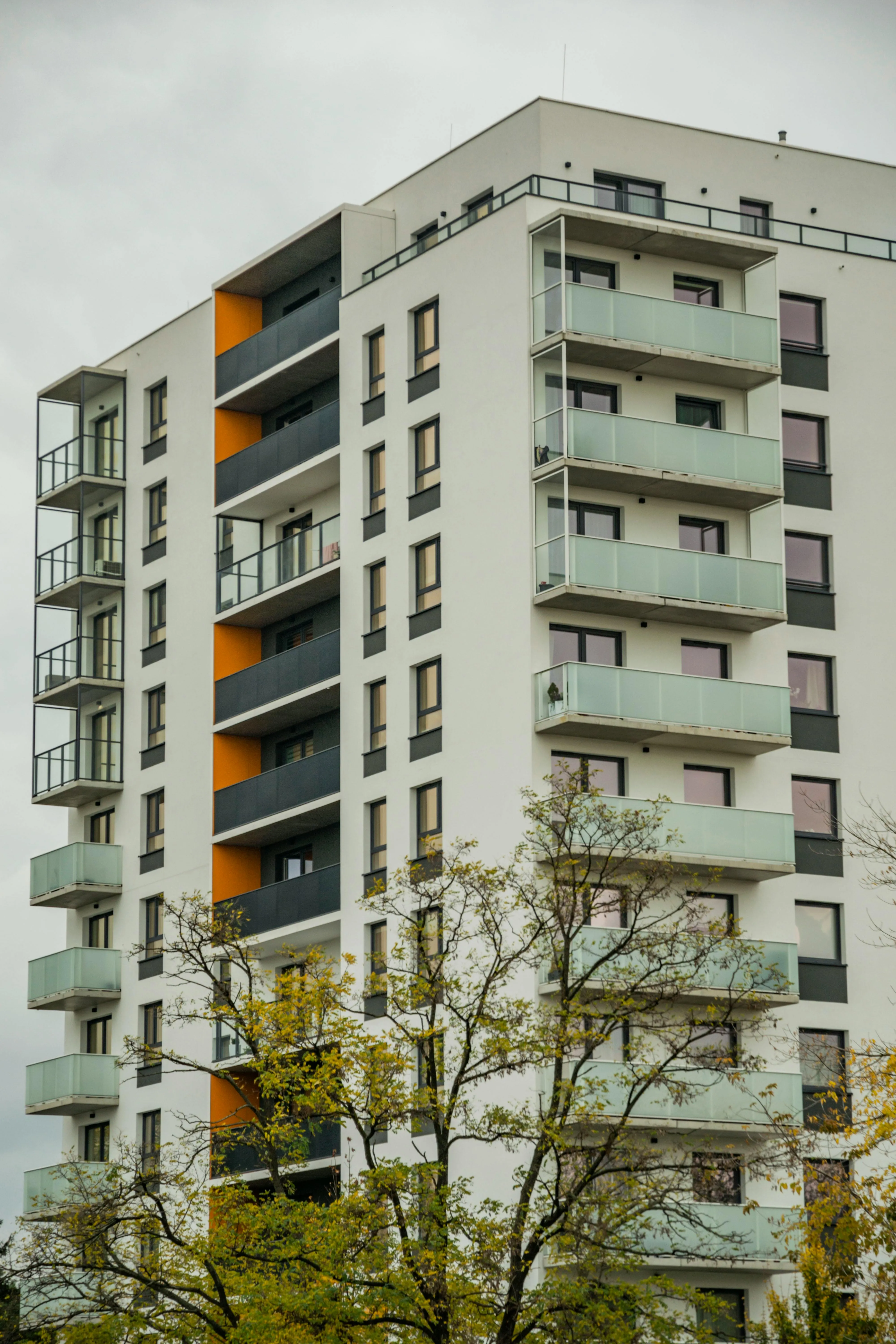 Modern multi-story apartment building with glass balconies and orange accent walls under cloudy sky.