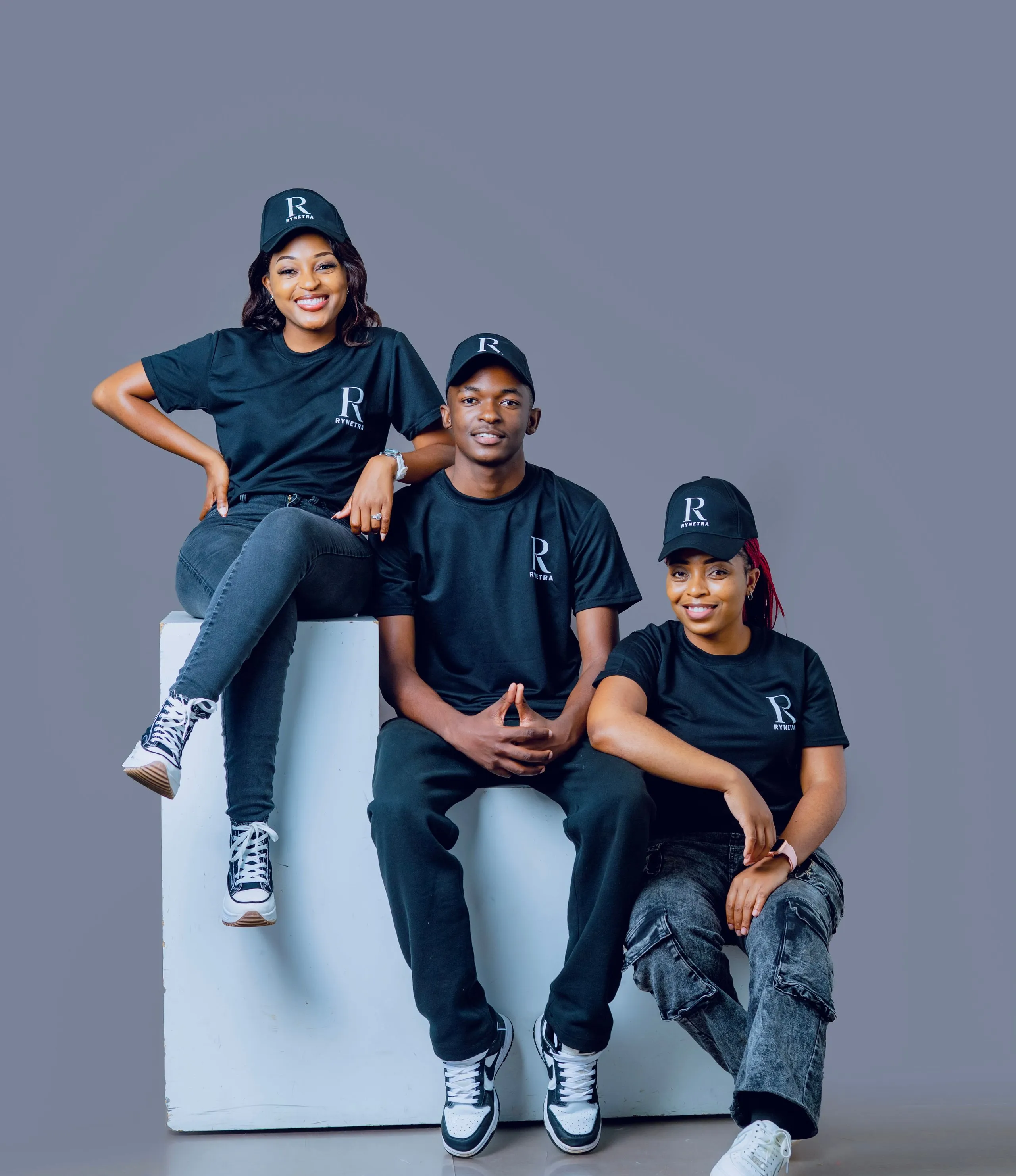Three people wearing matching black Rynetra shirts and caps posing against a gray background.