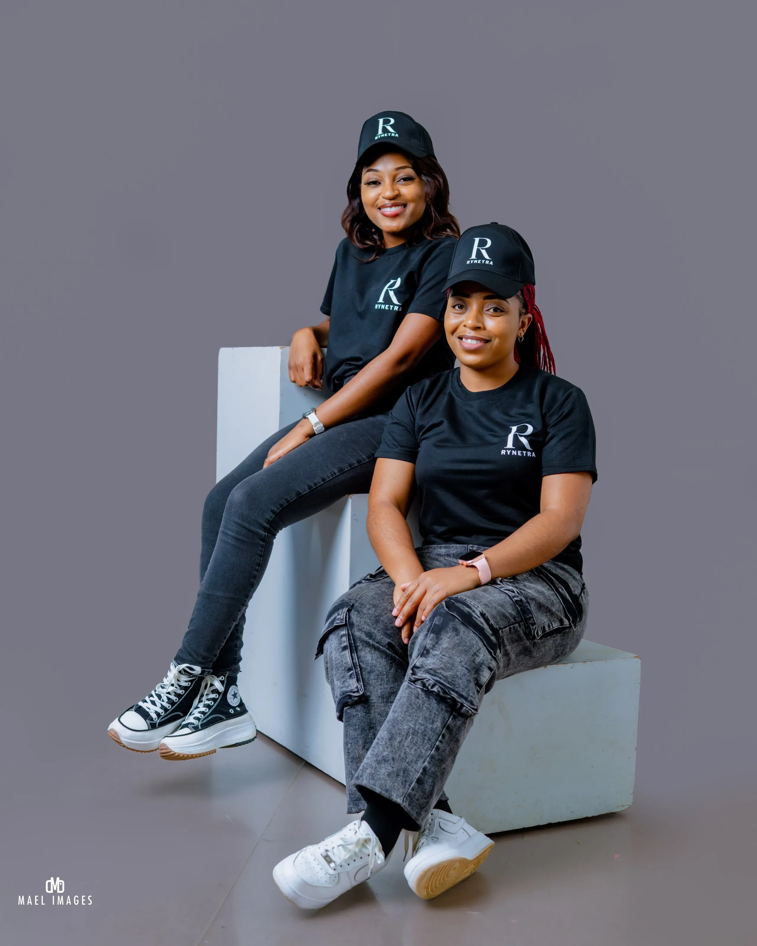 Two women smiling in black Rynetra shirts and caps sitting on white blocks against gray background.