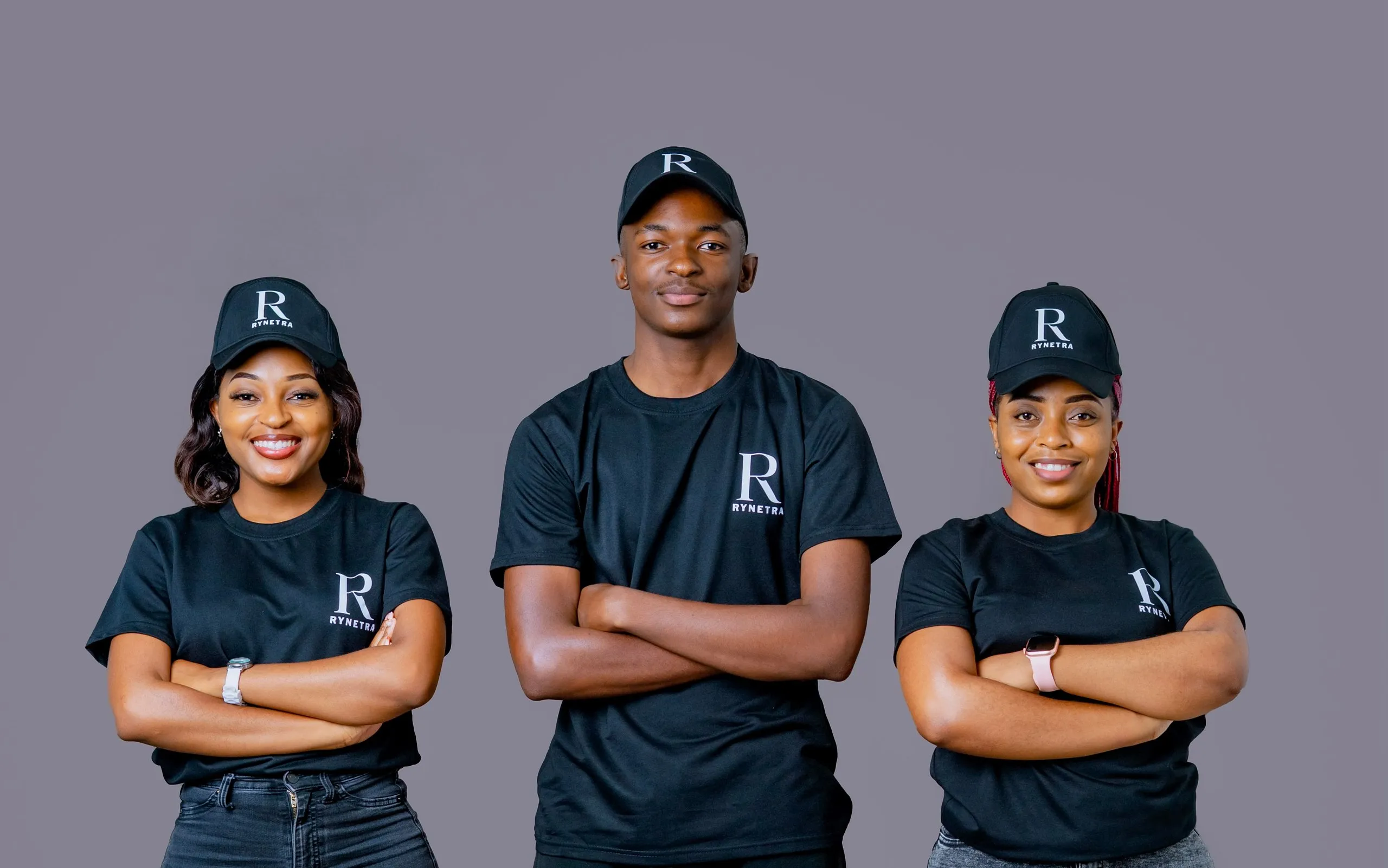 Three people wearing black Rynetra shirts and caps stand with arms crossed smiling against gray background.