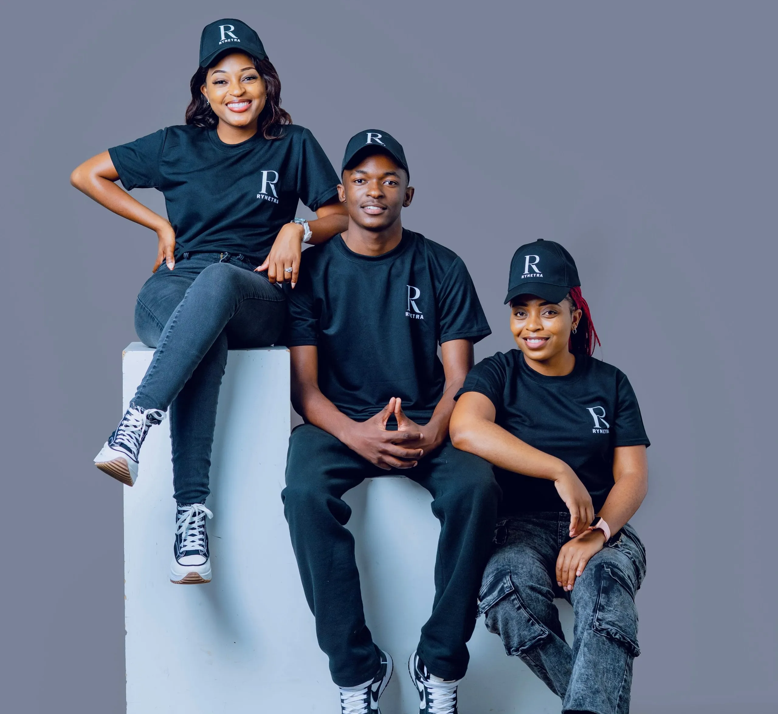 Three young adults wearing black Rynetra caps and shirts, smiling against a gray background.