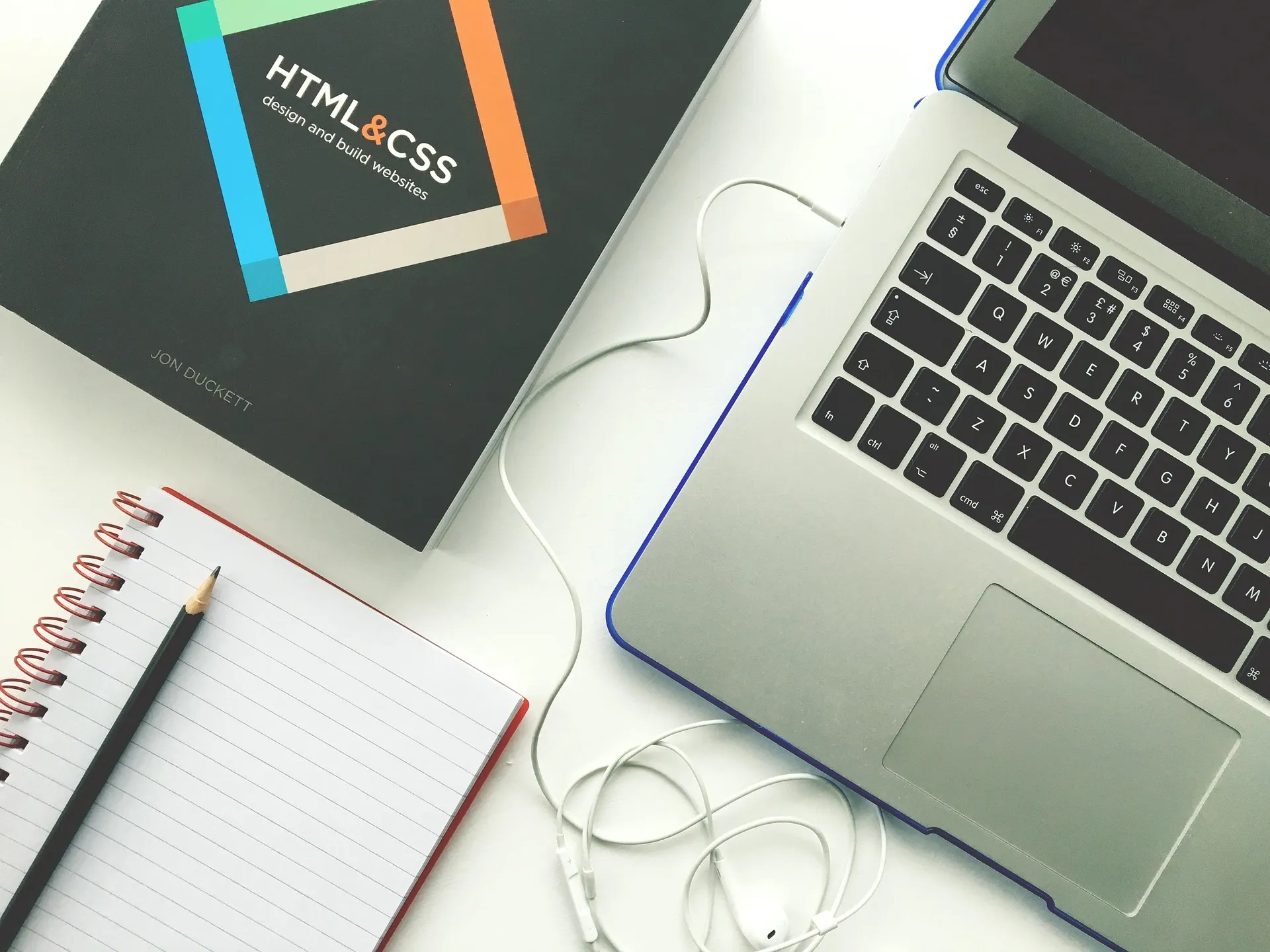 Laptop keyboard, notebook with pencil, HTML and CSS book, and earphones on white desk surface.